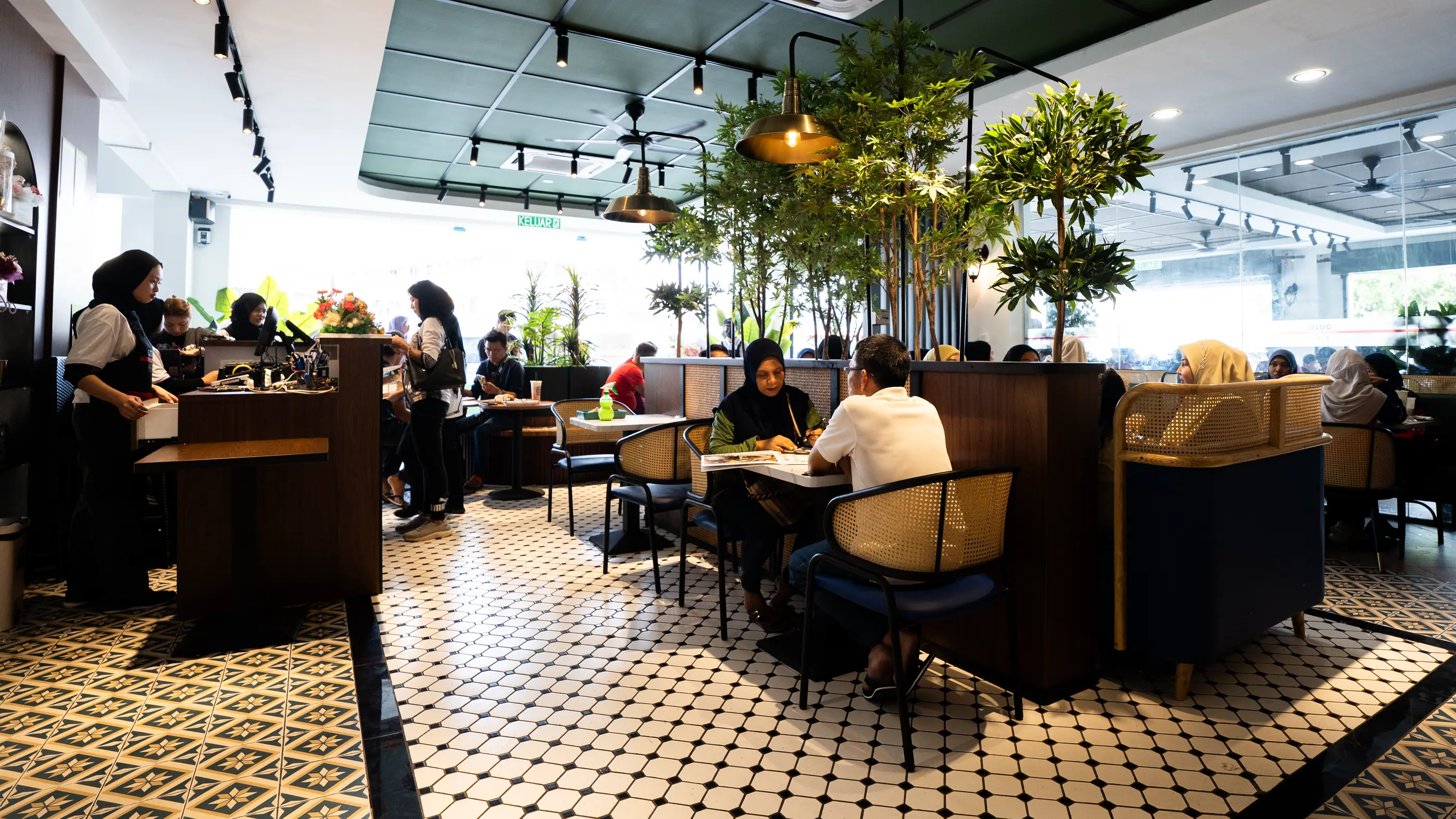 A busy DOJO kopi house: guests in rattan booths among indoor trees, patterned tiles and pendant lights