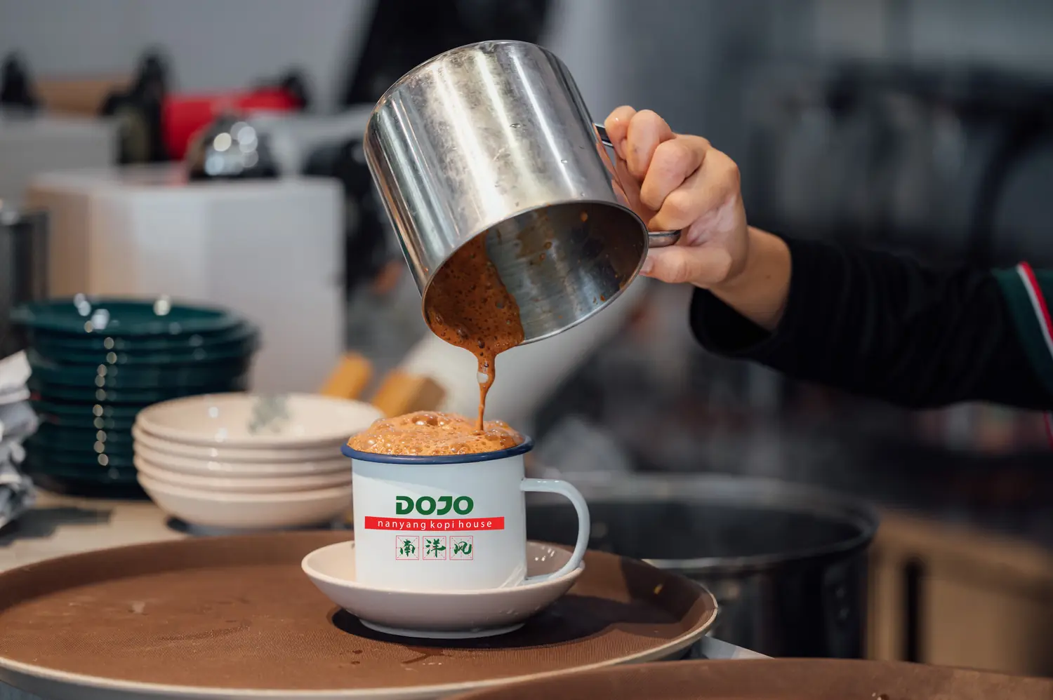A barista pours frothy kopi into a DOJO enamel mug