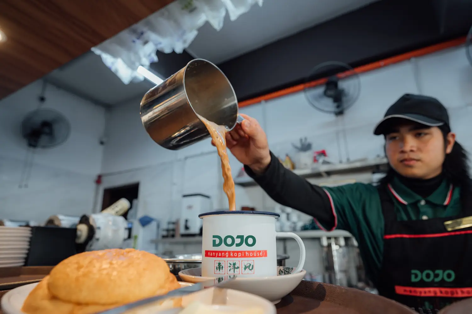 A DOJO barista pulls kopi into an enamel mug beside a golden polo bun